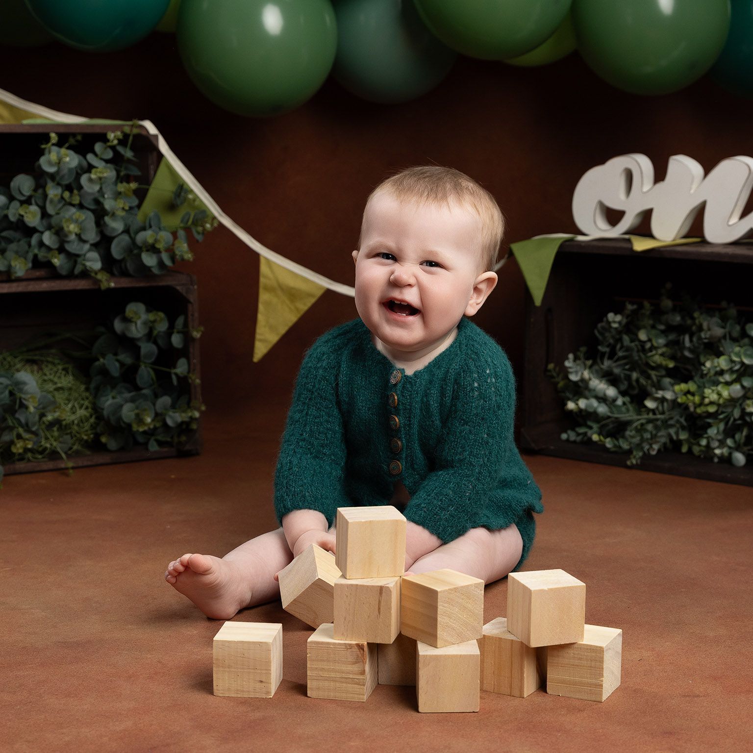 Happy baby in green sweater, playing with blocks, brown backdrop, green balloons.