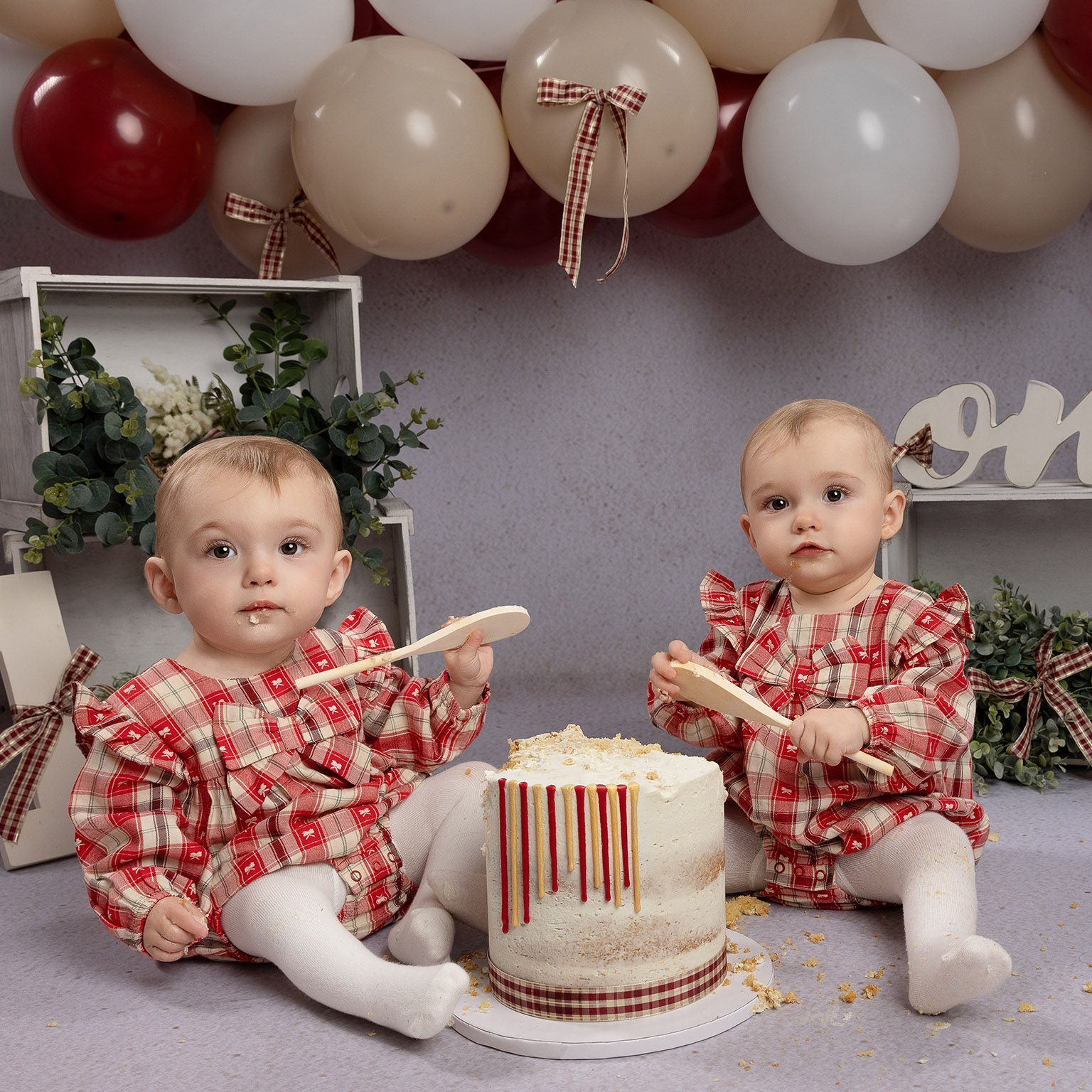 Twin babies in matching red plaid outfits, eating cake at their first birthday. Balloons overhead.