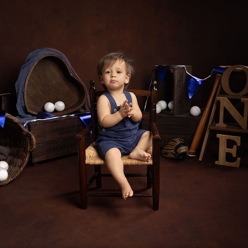 Baby in denim overalls sits in a chair, with baseball theme decor, brown background.