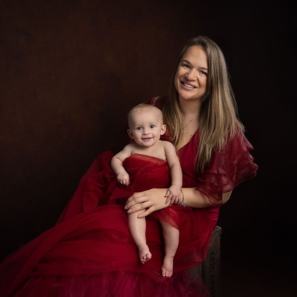 Woman in red dress holding smiling baby; brown background.