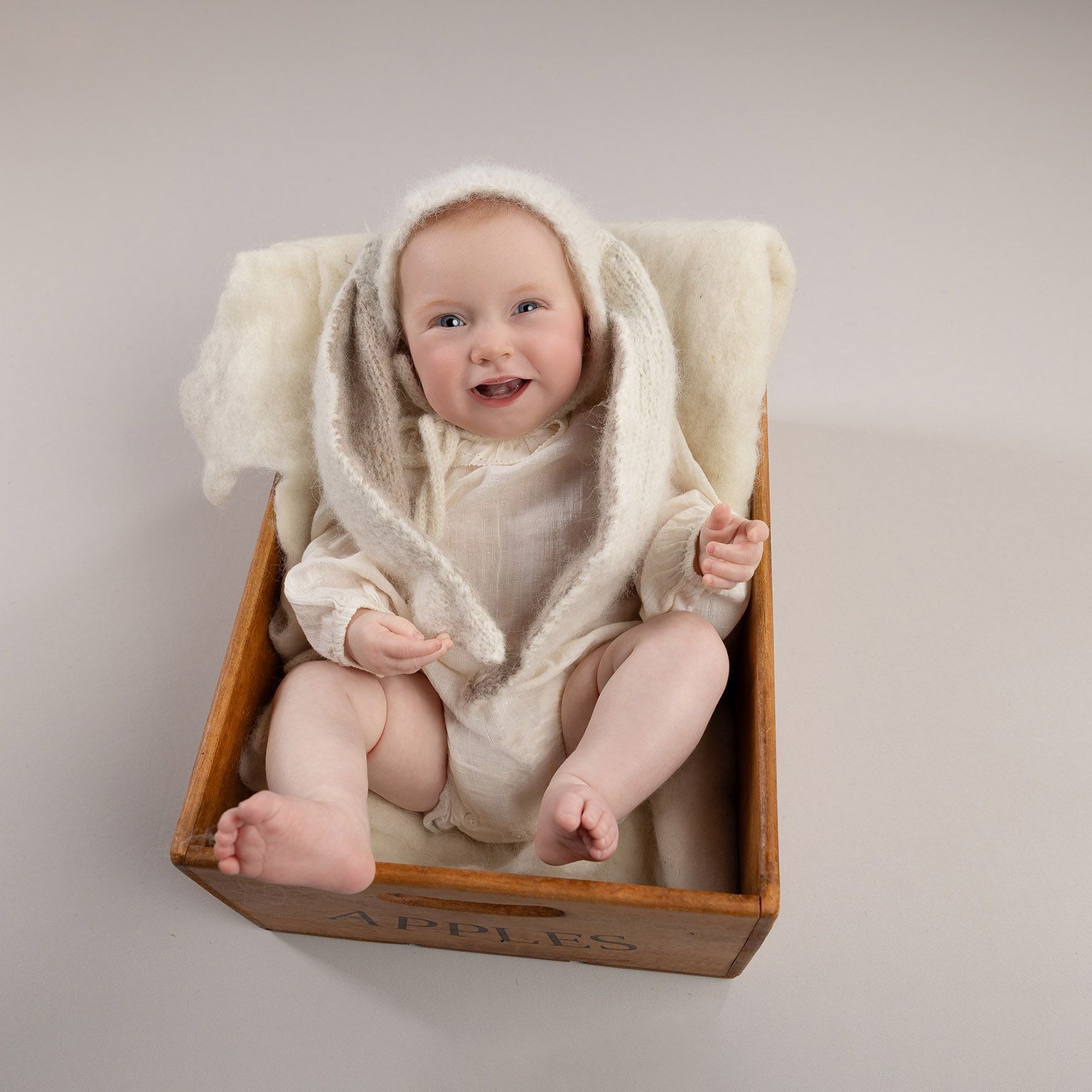 Baby wearing bunny outfit, sitting in a wooden box, smiling.
