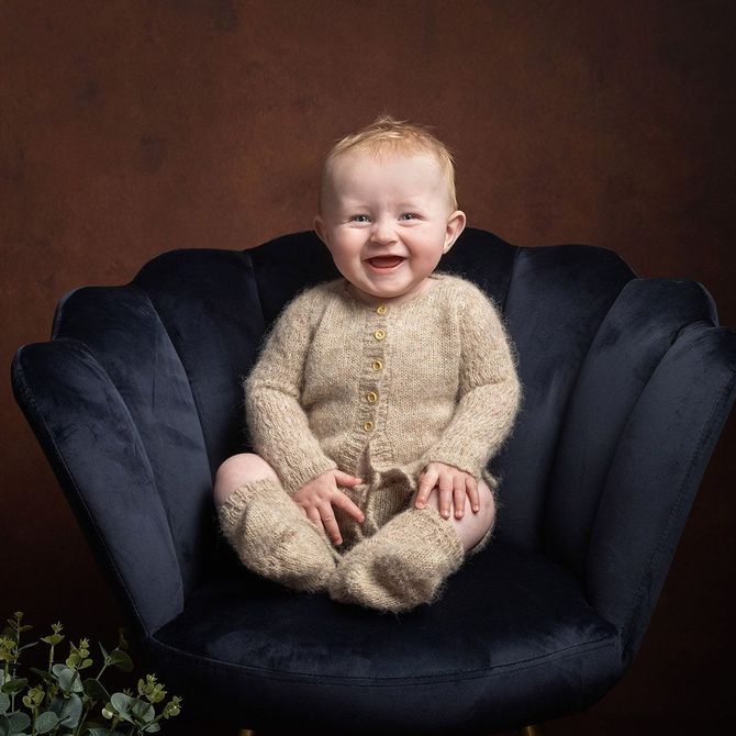 Smiling baby in a tan outfit sits in a navy blue velvet chair, brown backdrop.