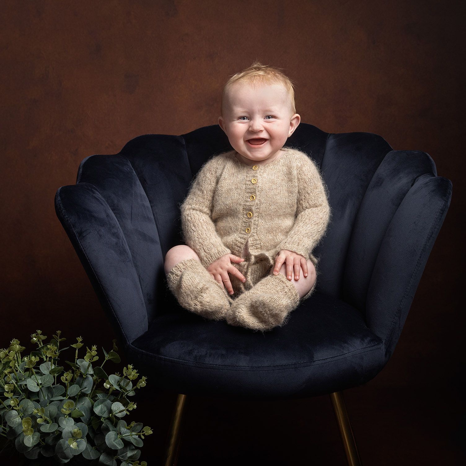Smiling baby in a fuzzy beige outfit sits in a dark blue velvet chair; brown backdrop; greenery in the foreground.