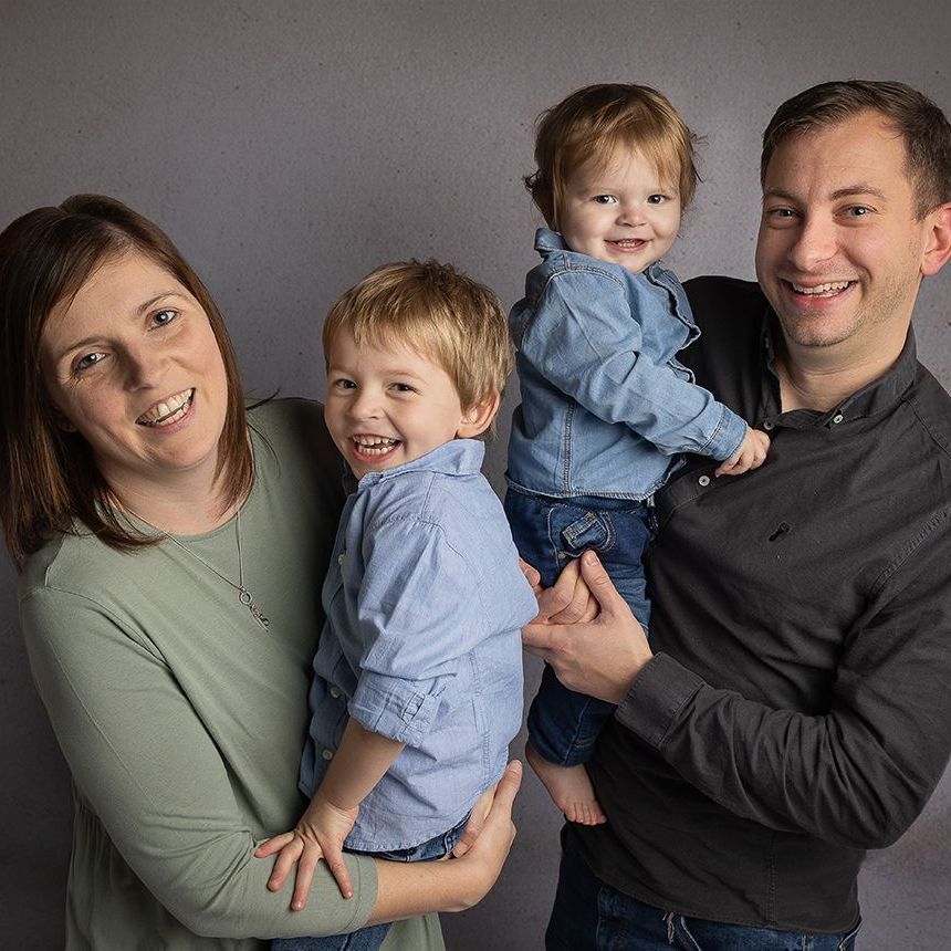 Family of four smiling, posing. Woman in green, man in black holding a child. Second child in blue.