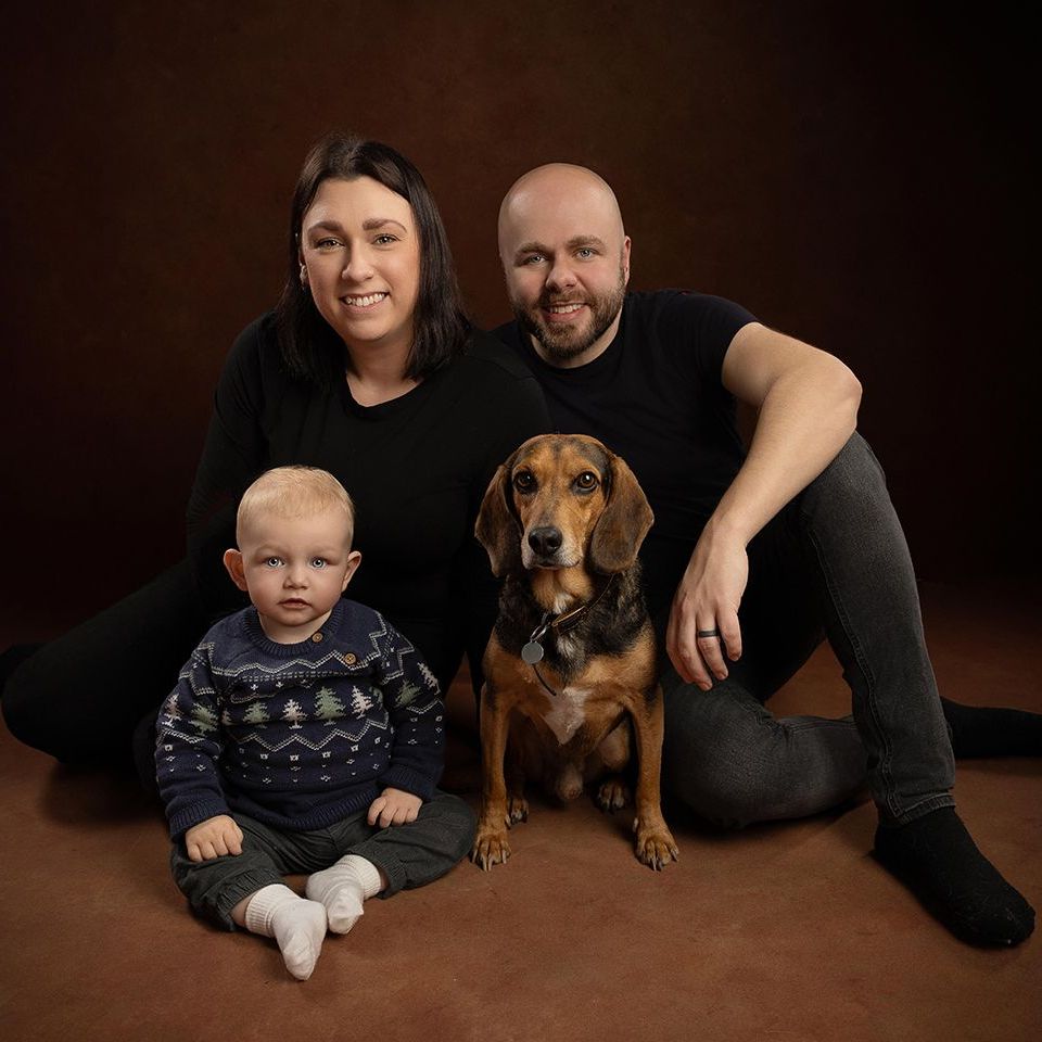 Family portrait: woman, man, baby, dog sit on brown surface, smiling at camera.