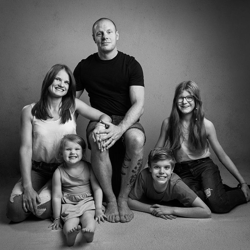 Family portrait: father seated, surrounded by kneeling wife and three children, all smiling. Black and white studio.