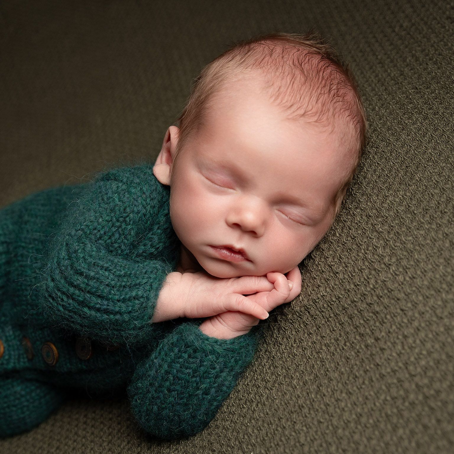 Newborn baby sleeping, wearing teal knit outfit, hands under chin, on olive blanket.