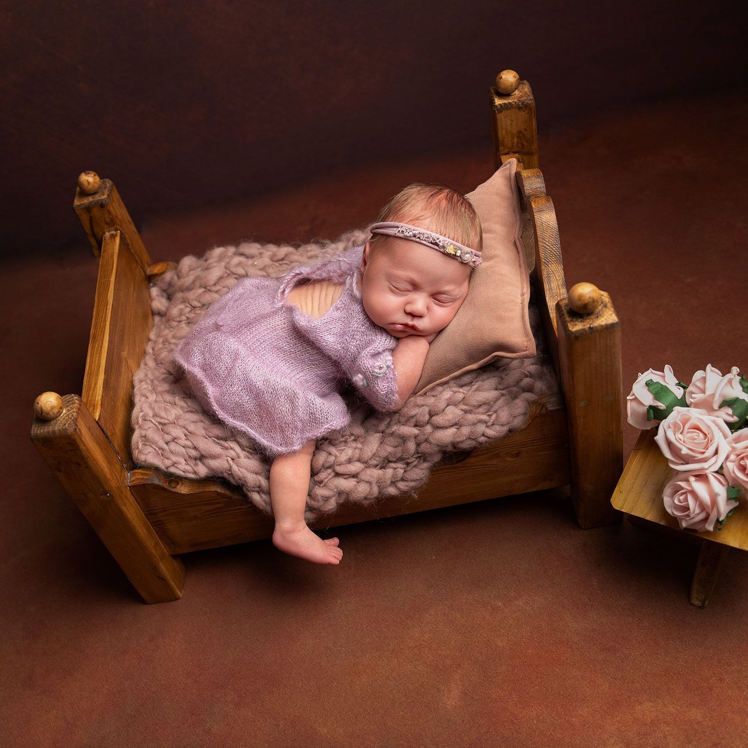 Newborn baby sleeping in a miniature wooden bed, wearing a knitted lavender dress and headband, brown background.