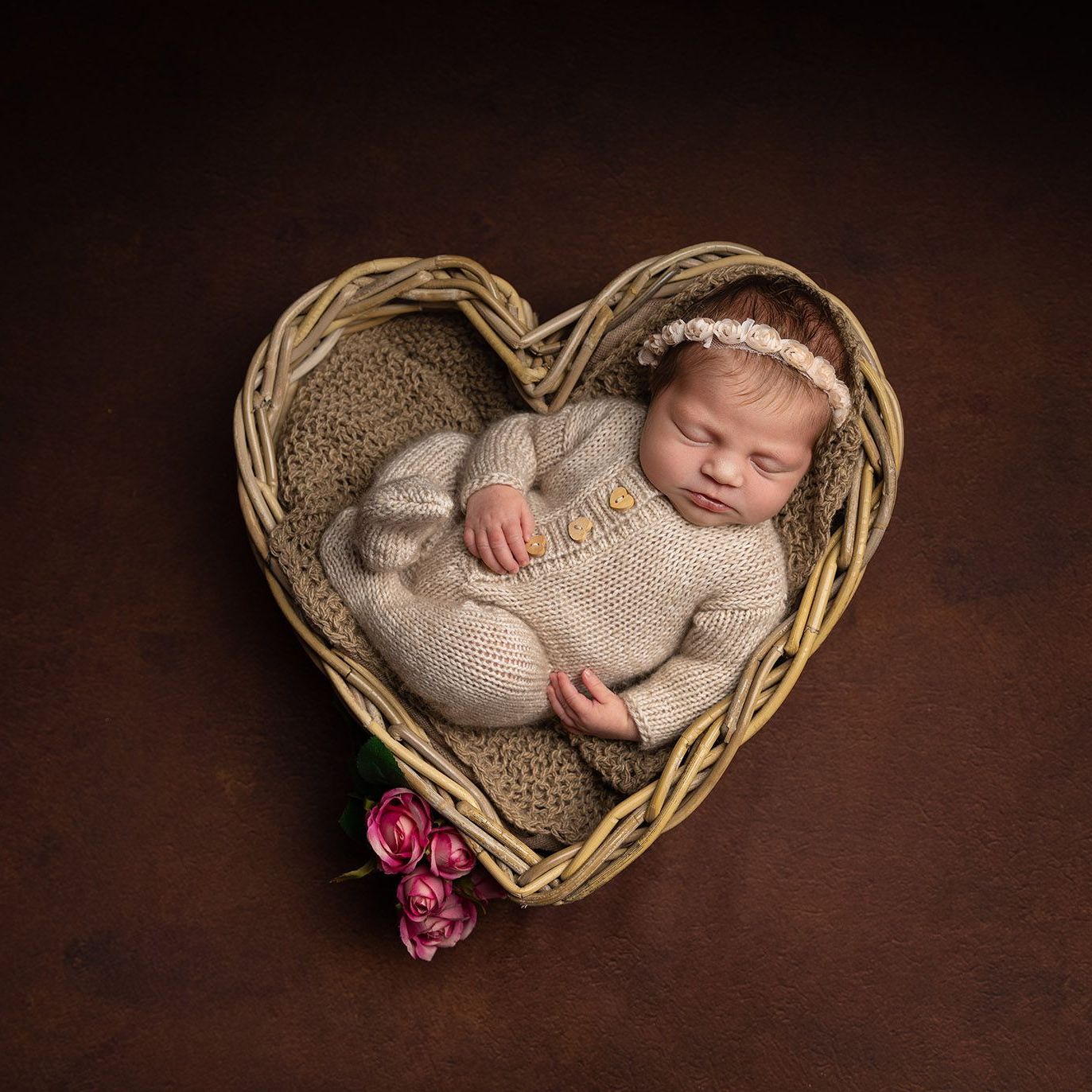 Newborn baby sleeping in a heart-shaped wicker basket, brown background, wearing a knitted outfit and headband.