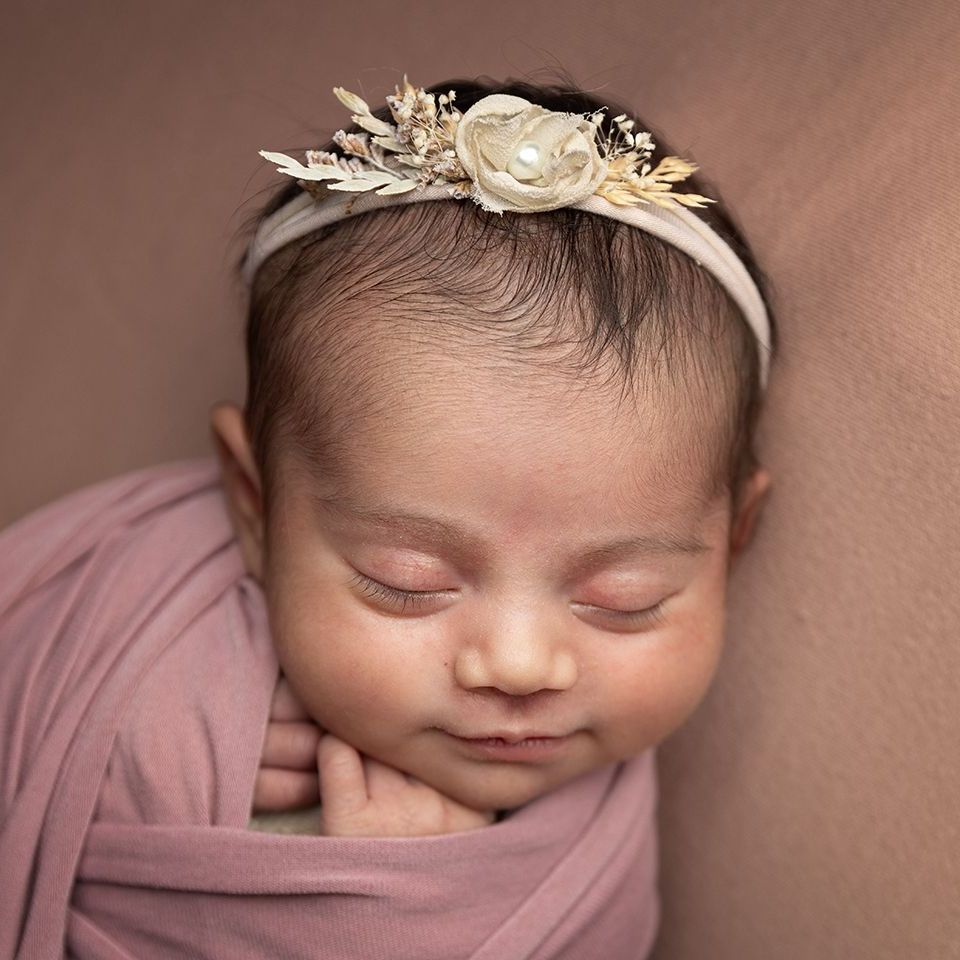 Newborn baby wrapped in pink, wearing floral headband, eyes closed.