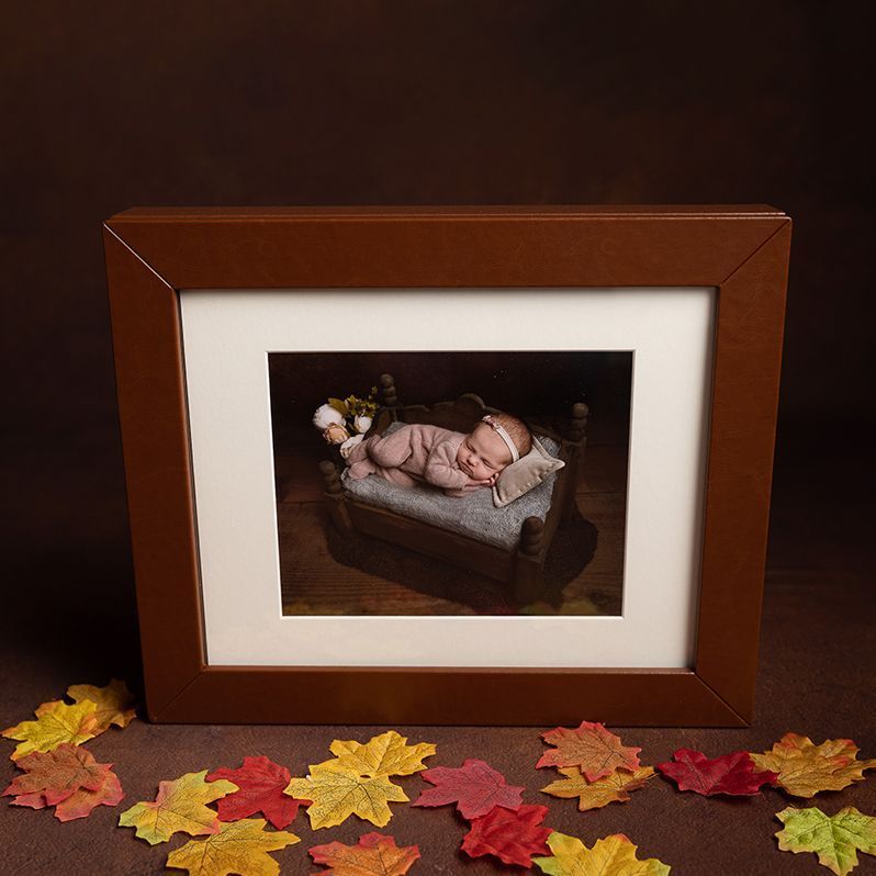 Newborn sleeps in a miniature bed framed by brown mat and frame, surrounded by colorful leaves.