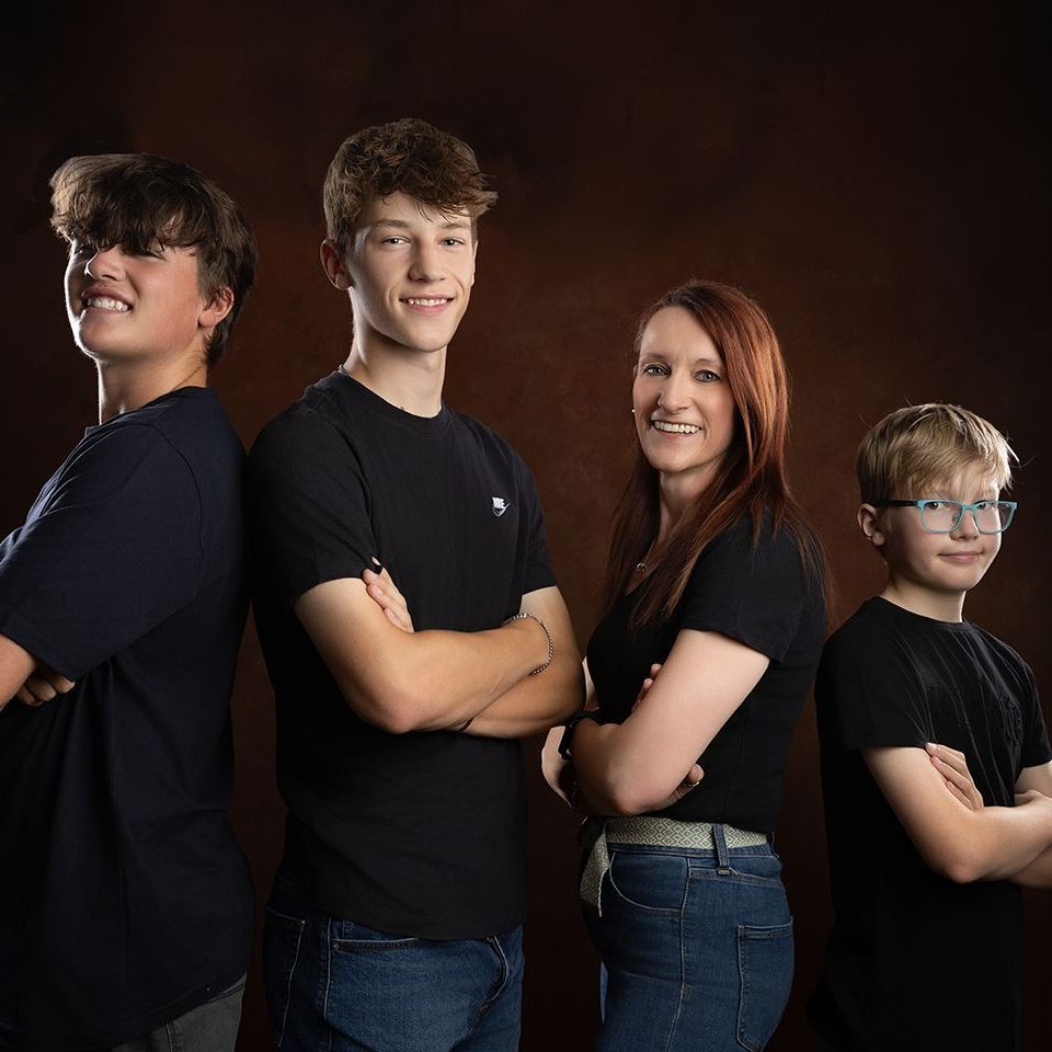 Family portrait; two teen boys, a woman, and a boy pose with arms crossed, smiling in front of a brown backdrop.
