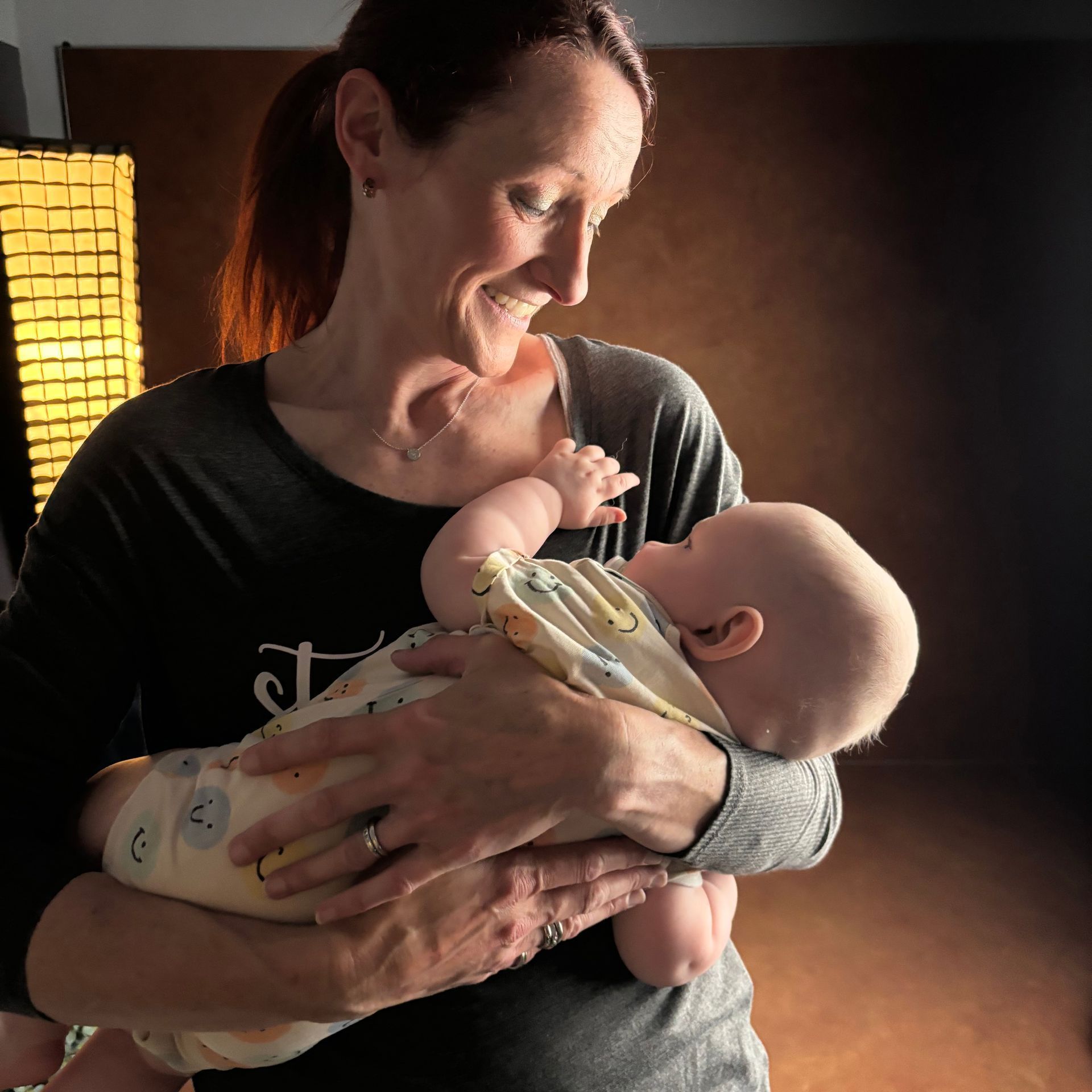 Woman with auburn hair smiles, holding a baby in a studio. Soft light, brown backdrop.