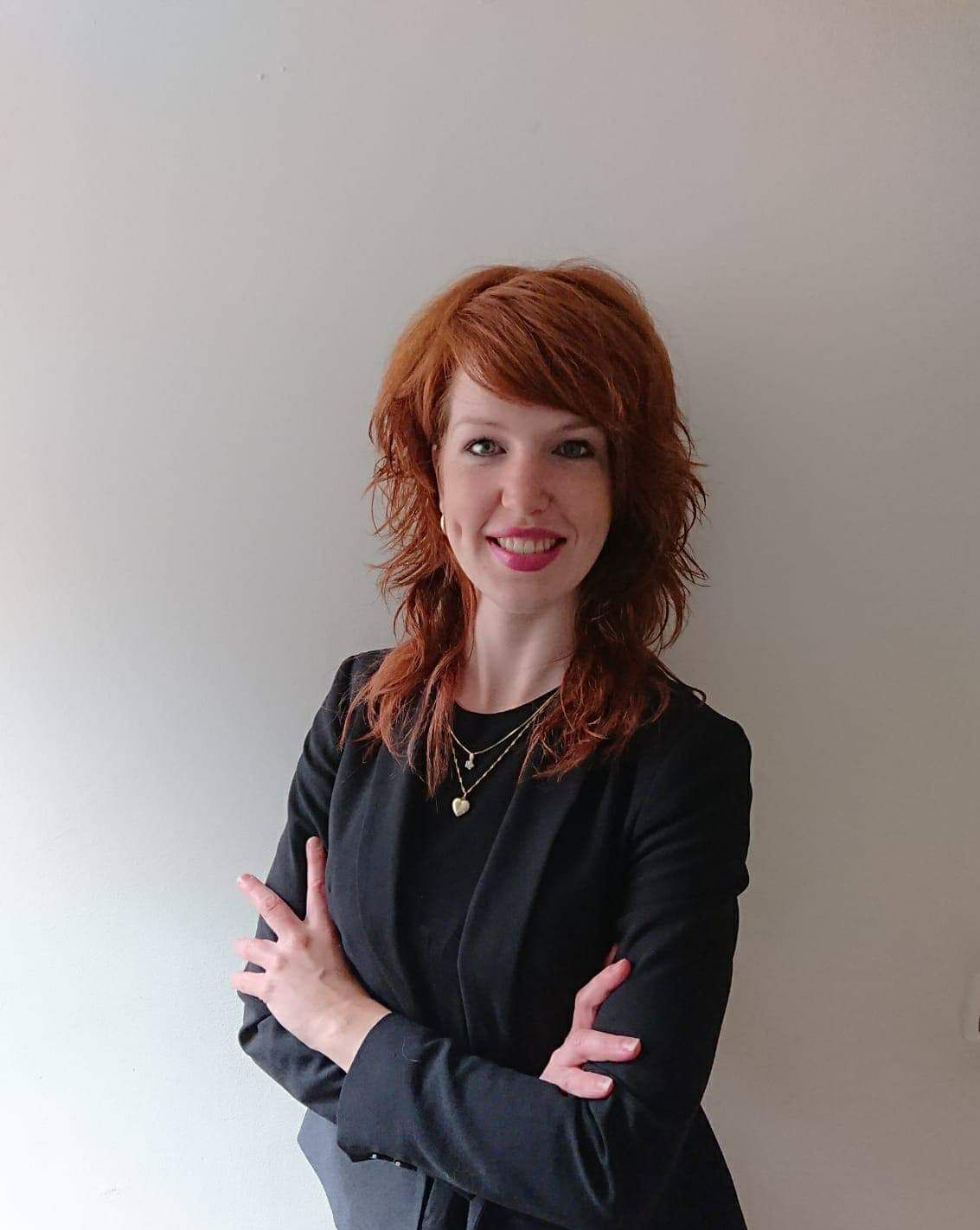 Vrouw met krullend rood haar, zwarte top, lachend met de armen over elkaar tegen een effen muur.