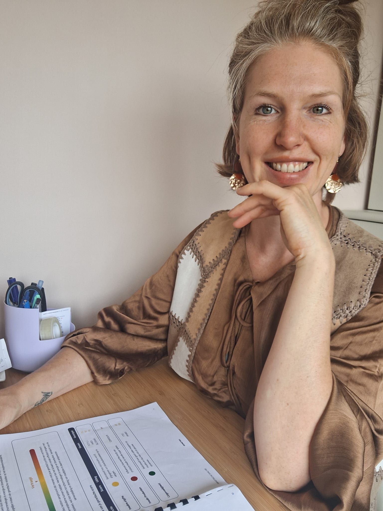 Een vrouw glimlacht achter een bureau met papieren en een pennenhouder, terwijl ze haar kin op haar hand laat rusten.