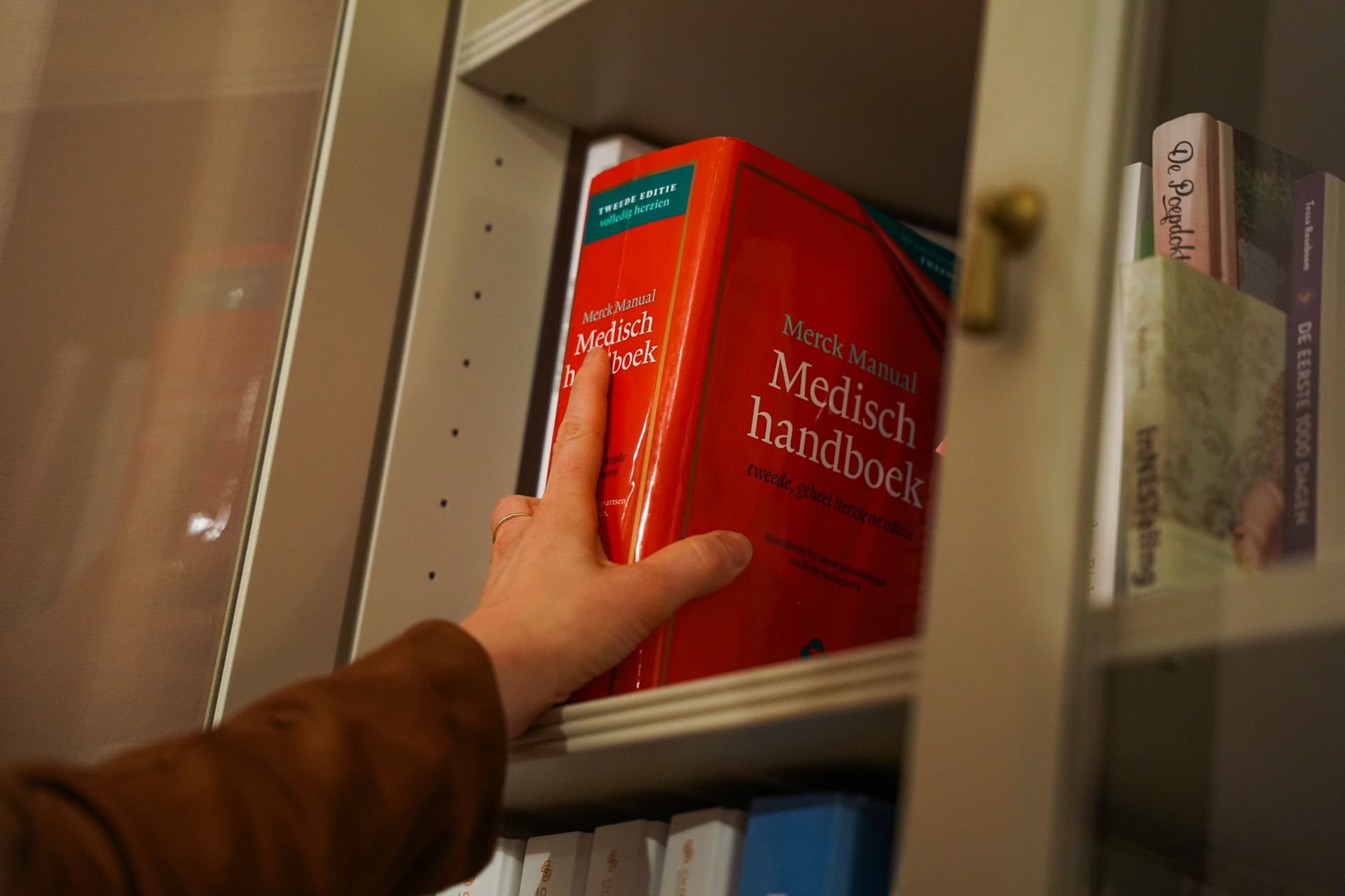 Een hand reikt naar een rood medisch handboek op een boekenplank achter glas.