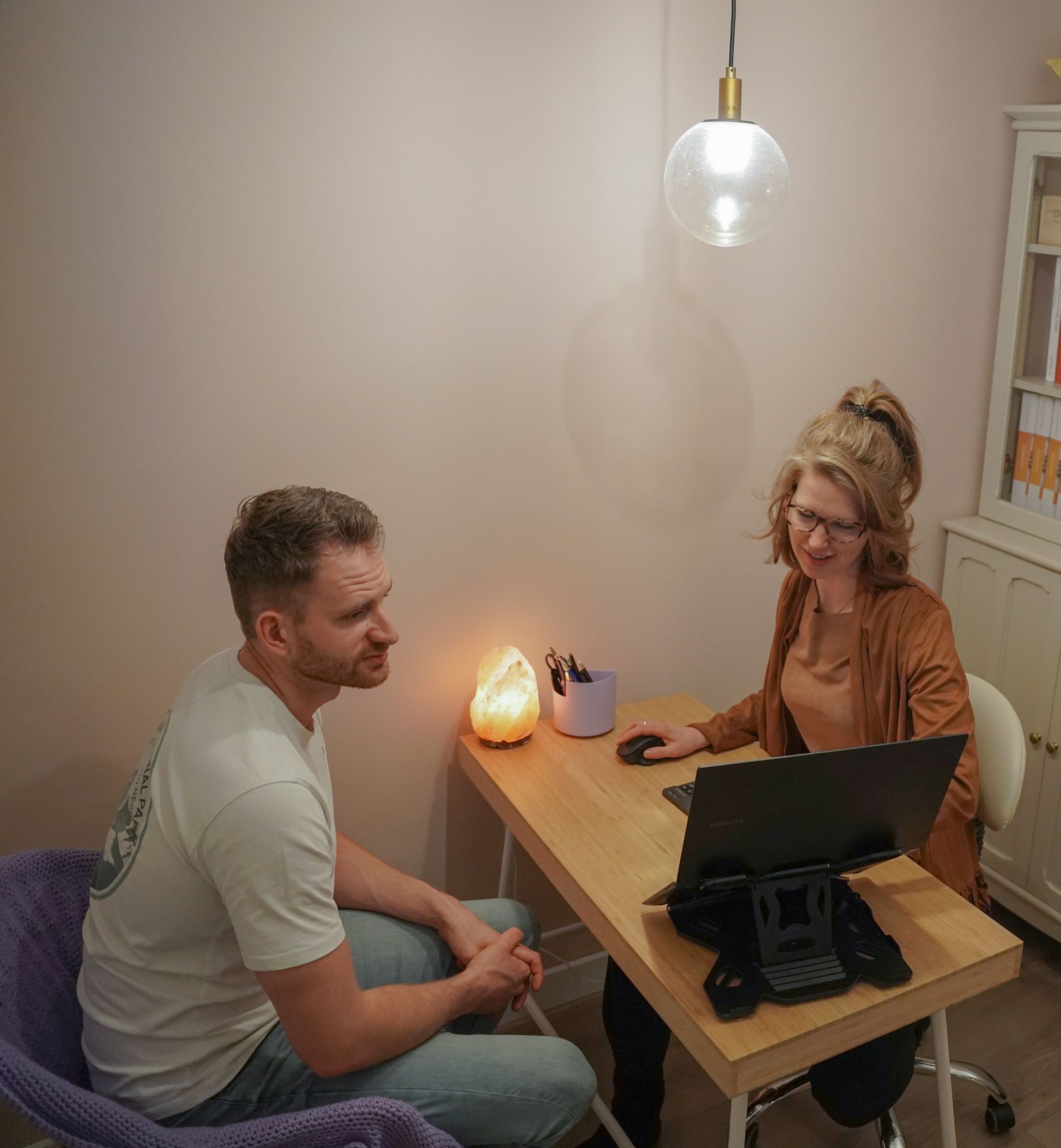 Twee mensen praten aan een klein bureau met een laptop in een zacht verlichte kamer.