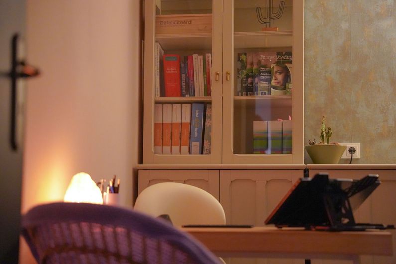 Een knusse kamer met een bureau, een witte stoel, een brandende lamp en een glazen vitrinekast op de achtergrond.