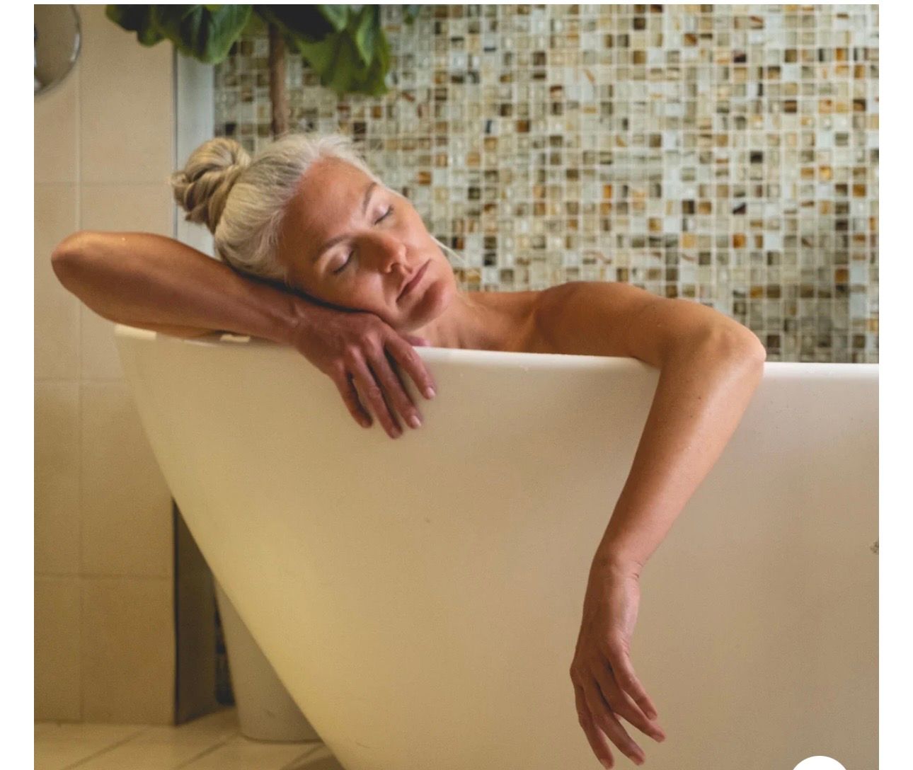 Person relaxing in a bathtub, leaning over the side with eyes closed, in a tiled bathroom.