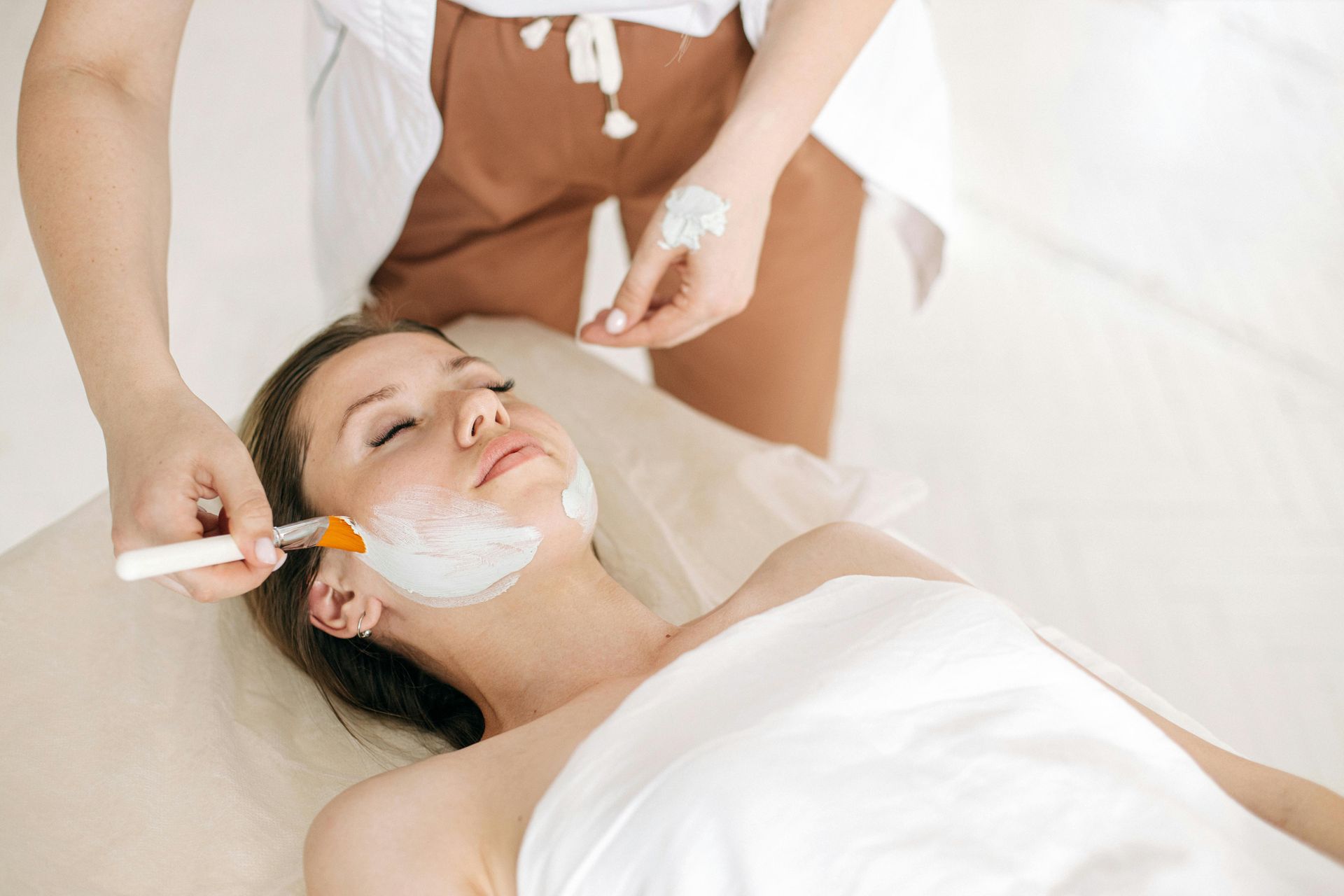 Woman receiving a facial treatment. A beautician applies a white mask to her face in a spa setting.