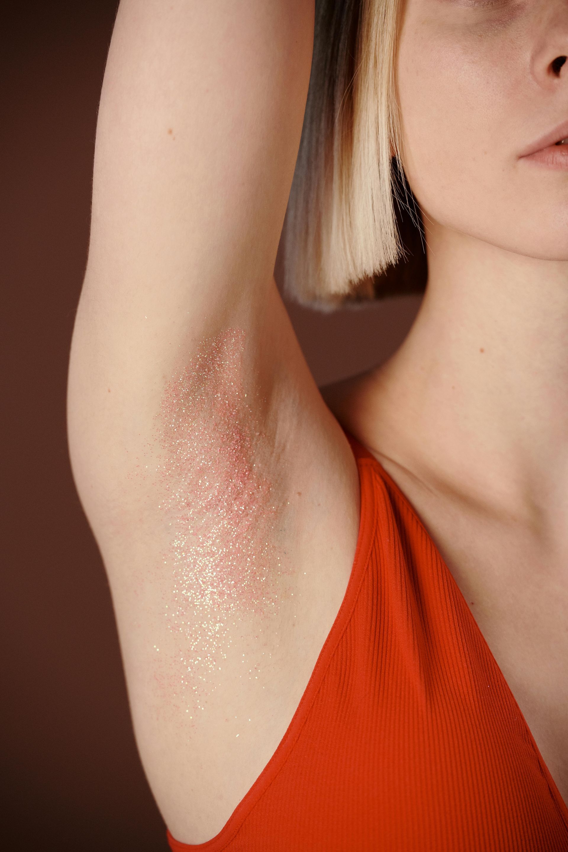 Woman's arm with glitter-covered armpit, wearing a red top, against a brown background.