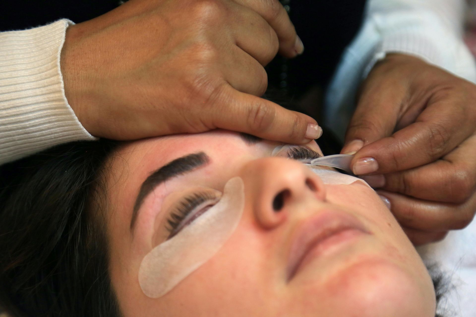 A woman getting eyelash extensions applied. Technician's hands are at her face, working on upper lashes.