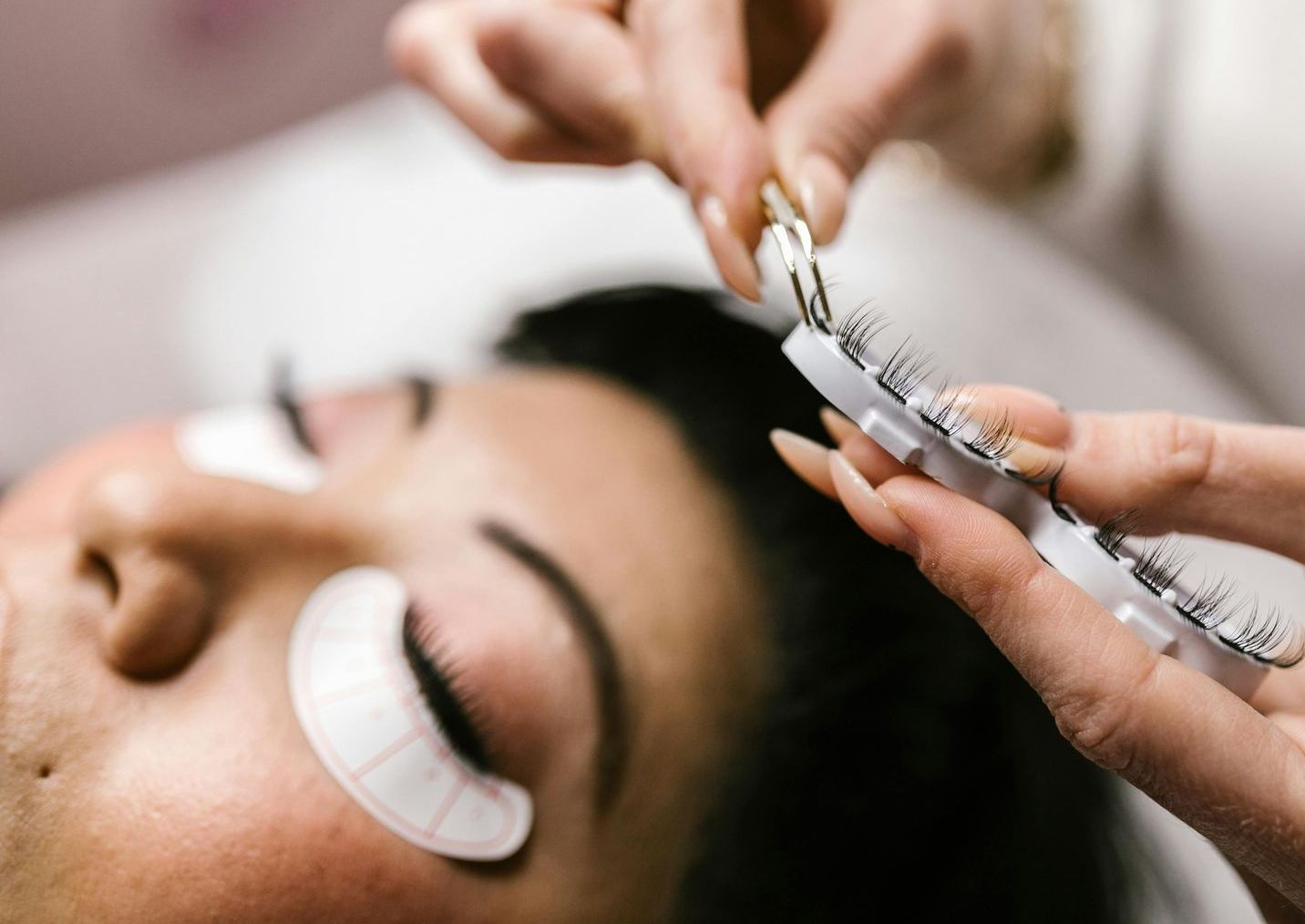 Woman receiving eyelash extensions in a salon; technician holding lashes and tools.
