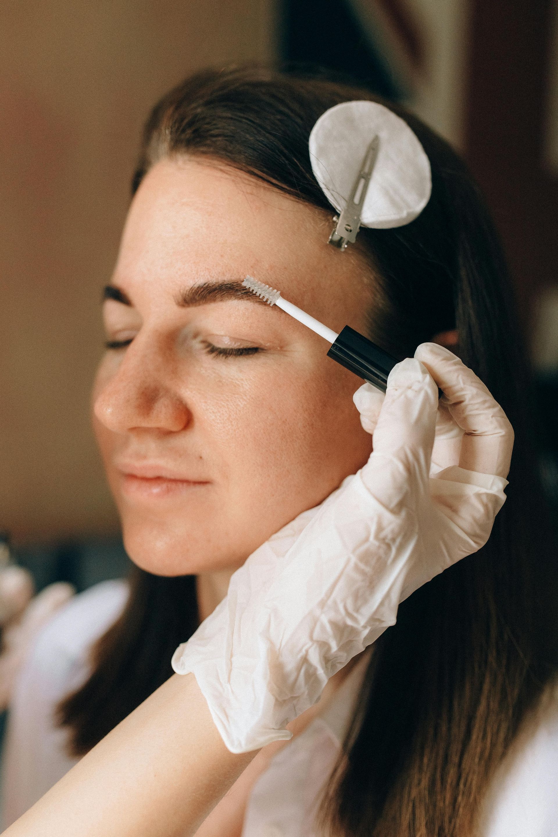Woman getting eyebrow makeup applied with her eyes closed, wearing gloves.