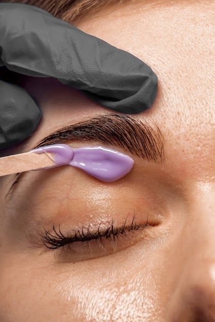 Close-up: A person's eyebrow being waxed. Purple wax applied with a spatula by a gloved hand. Eyes are closed.