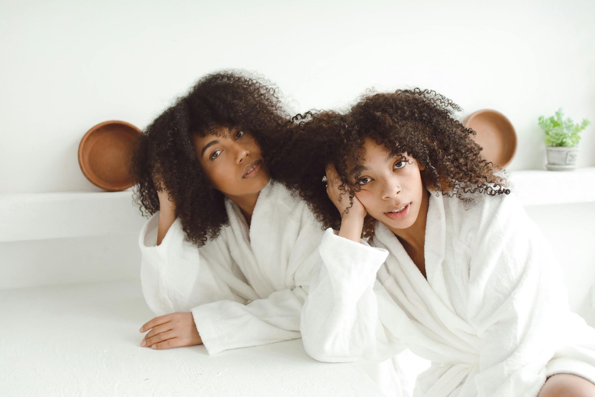 Two women in white robes leaning on white surface; curly hair; light-skinned; serene expressions.