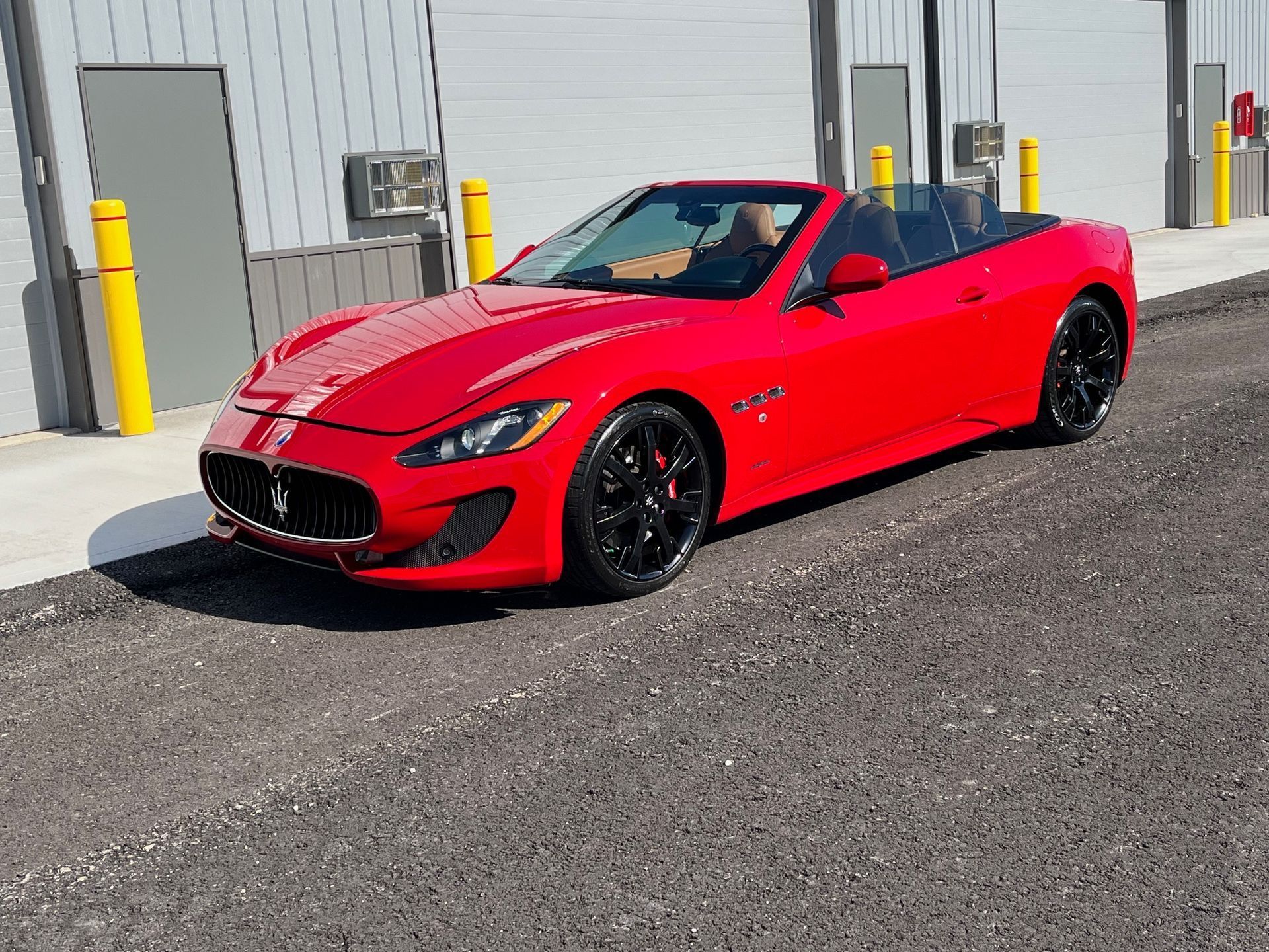 Red sports car parked outside a warehouse on asphalt, with a gray building and yellow bollards behind it