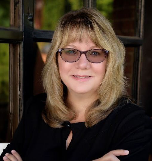 A woman wearing glasses and a black shirt is standing in front of a window.