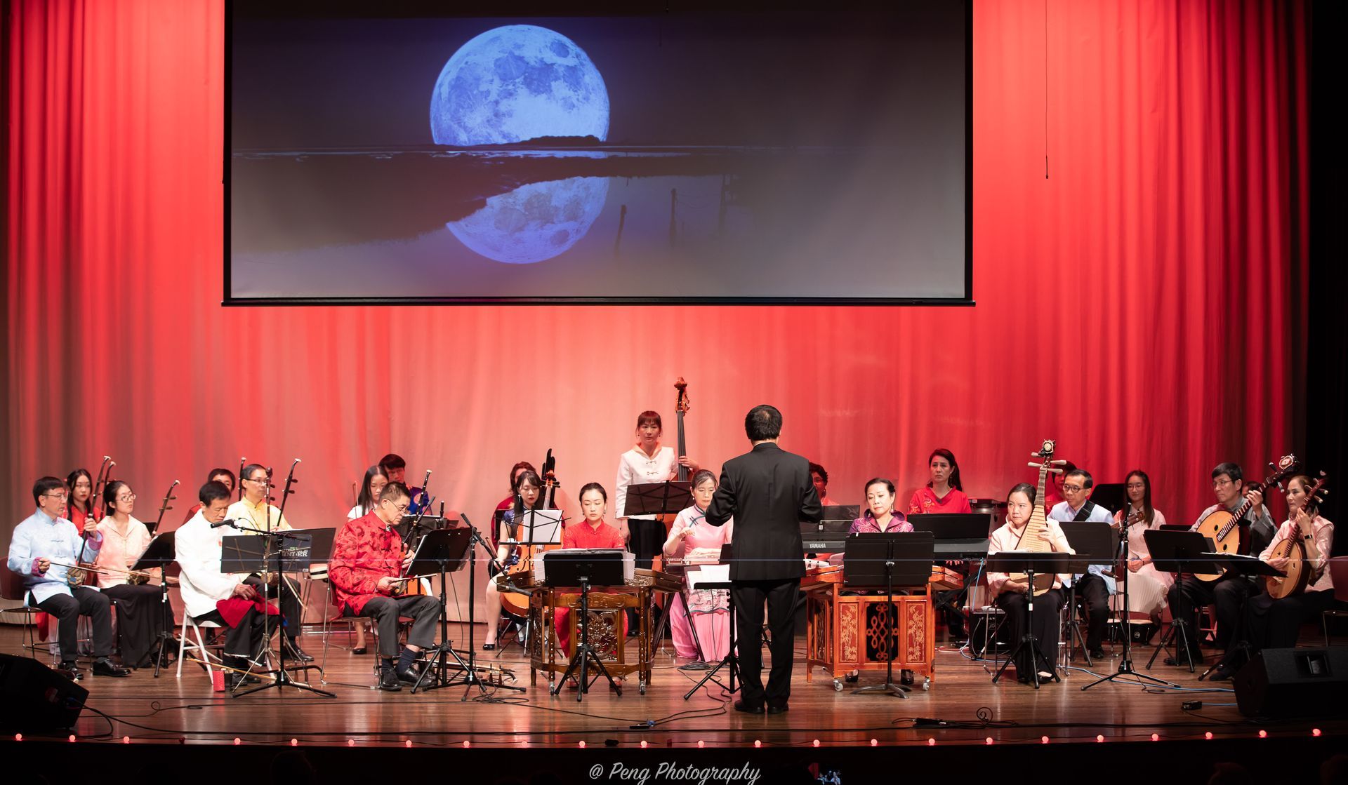 A man is leading an orchestra on a stage with a large screen behind them.