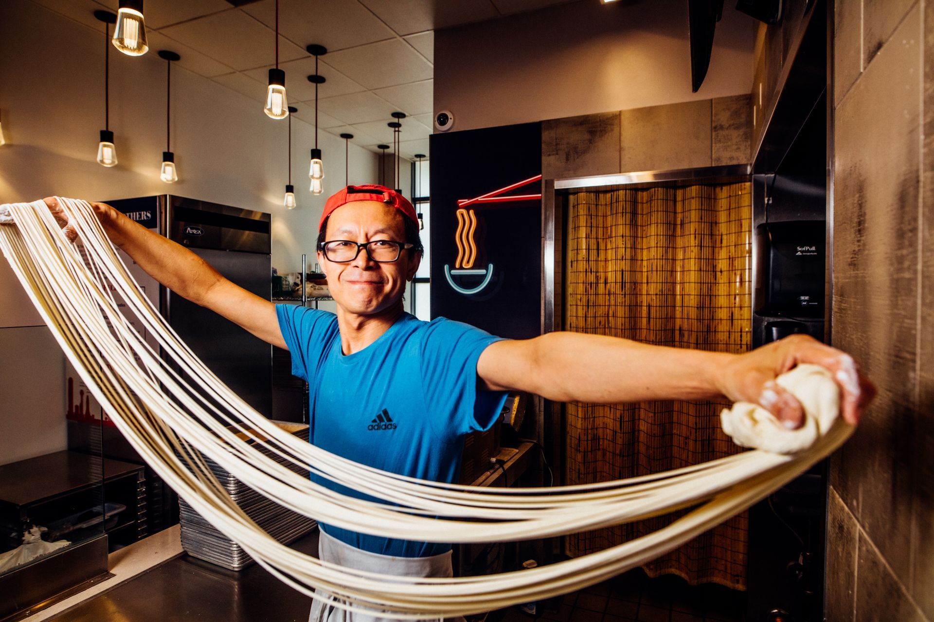 A man is making noodles in a kitchen with his arms outstretched.