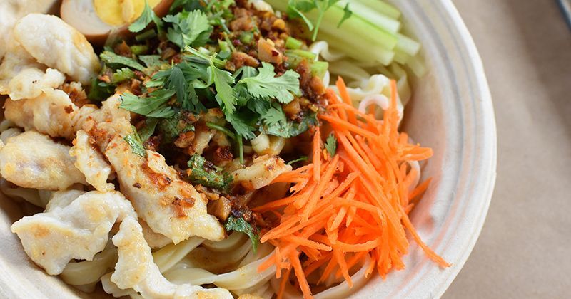 A bowl of food with noodles , carrots , celery and eggs on a table.
