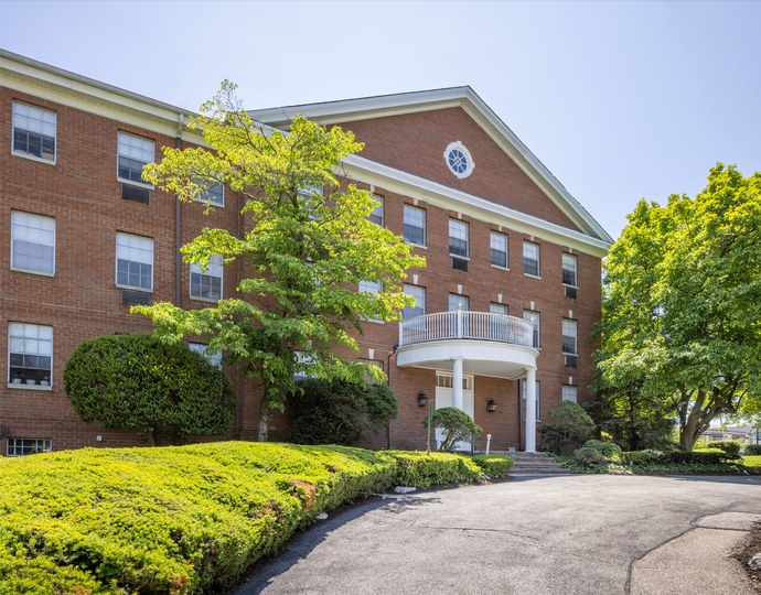 Red brick three-story building with white balcony and dormer window, set in a sunny day with green landscaping.