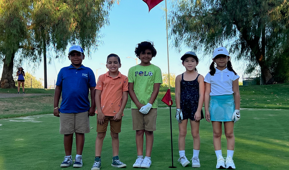 Five children standing on grass in a park, holding golf clubs, with trees and a red flag in the background.