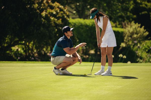 Golfer crouches beside woman on green, pointing at her putt on a sunny course Golfer crouches beside woman on green, pointing at her putt on a sunny course