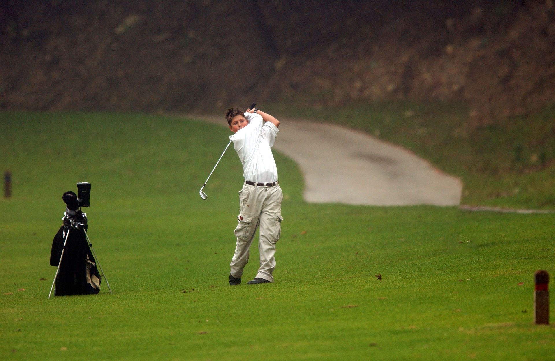 Golfer mid-swing on a green fairway, with a golf bag beside him and a winding path in the background Golfer mid-swing on a green fairway, with a golf bag beside him and a winding path in the background