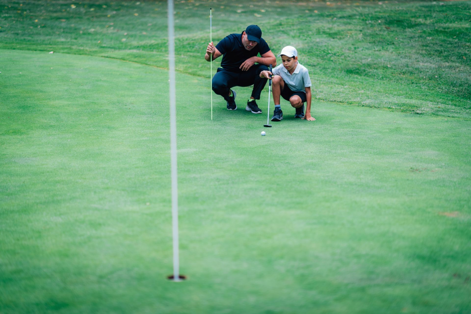 Two golfers crouch near a hole on a green putting surface, with the flagstick in the foreground. Two golfers crouch near a hole on a green putting surface, with the flagstick in the foreground.