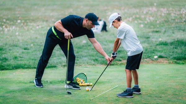 Two people practicing golf swing on a green, one instructor adjusting a child’s stance. Two people practicing golf swing on a green, one instructor adjusting a child’s stance.