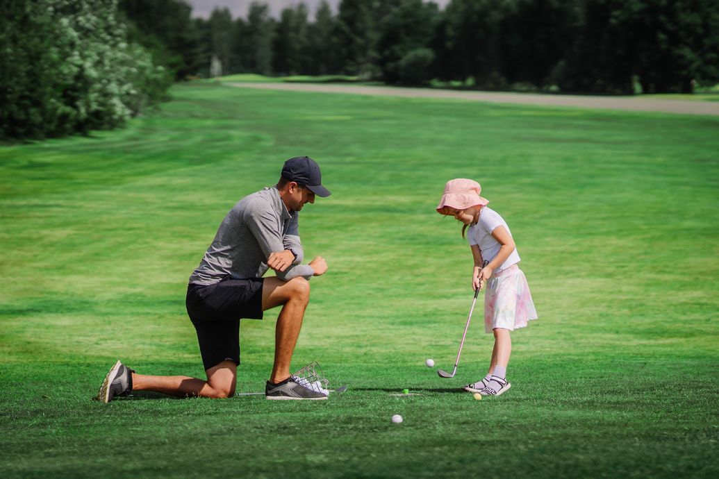 Golfer kneels beside child on green, coaching a putt on a golf course Golfer kneels beside child on green, coaching a putt on a golf course