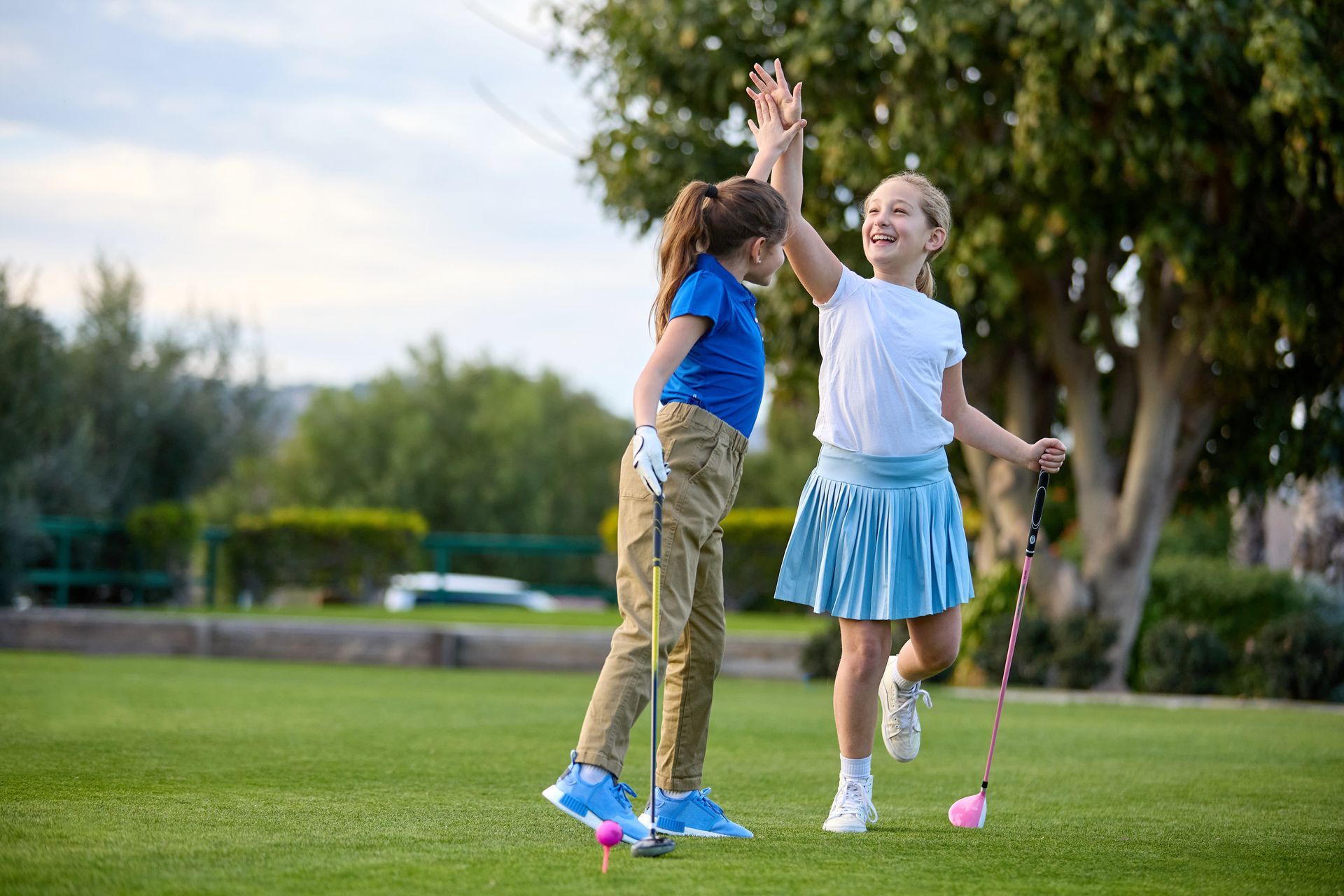 Two children high-five on a golf course, one holding a pink golf club.