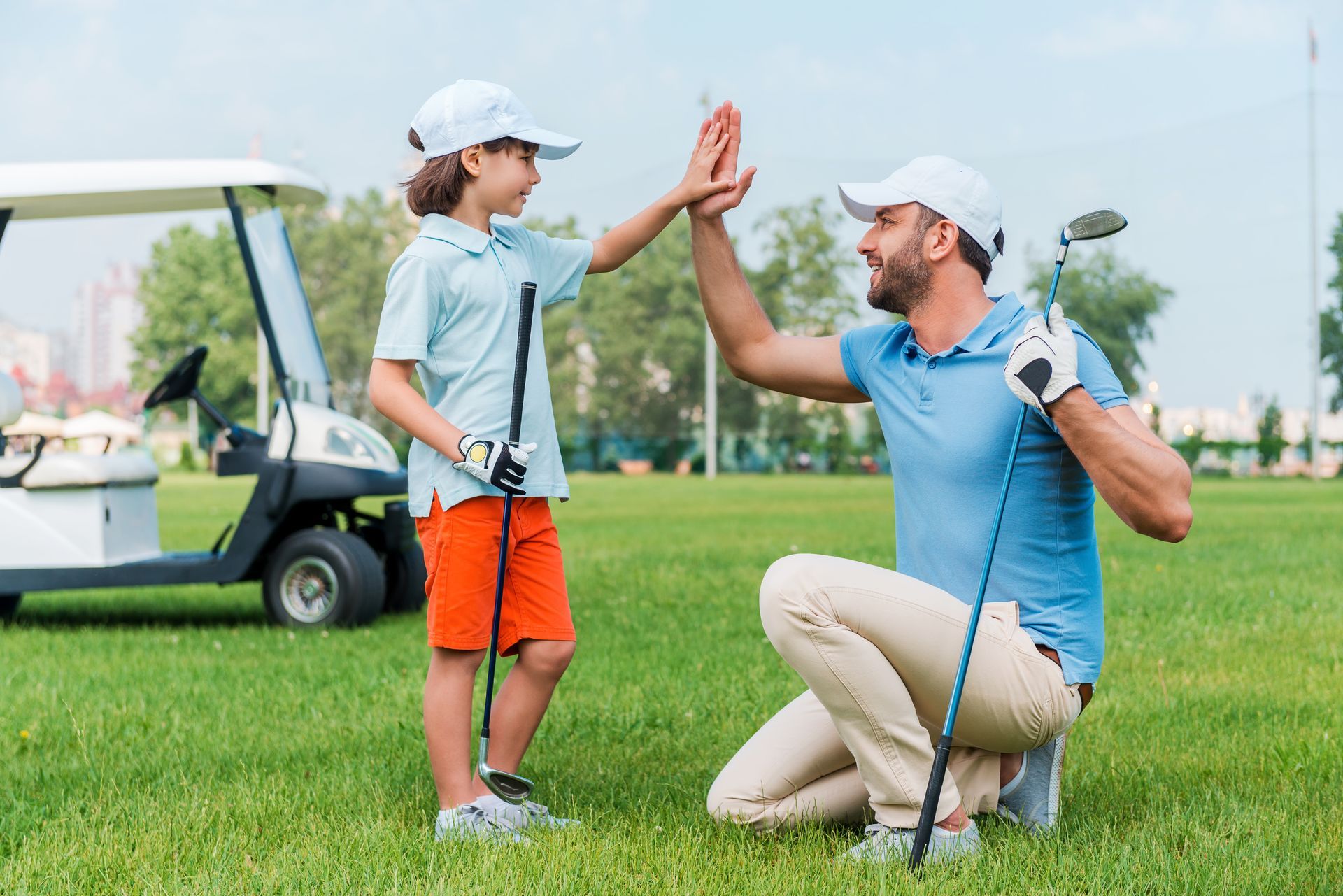 Golfer kneeling to high-five a child on a golf course, with a golf cart in the background Golfer kneeling to high-five a child on a golf course, with a golf cart in the background