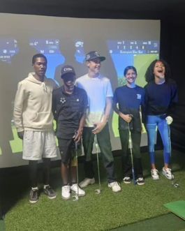 Five people posing with golf clubs at an indoor golf simulator, smiling and standing on turf