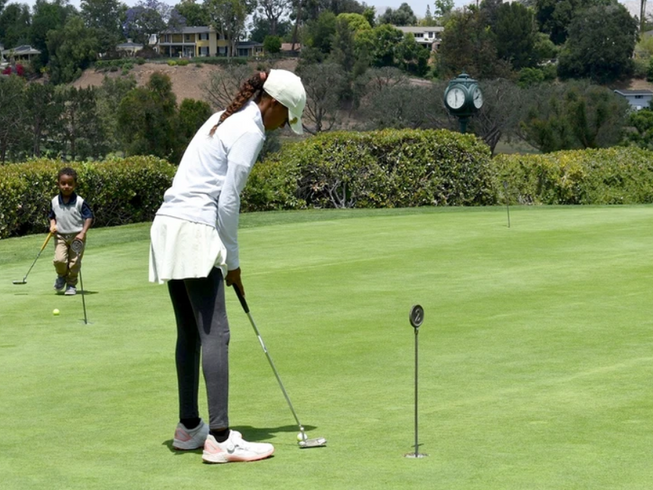 Golfer lining up a putt on a green, with a child nearby and trees in the background