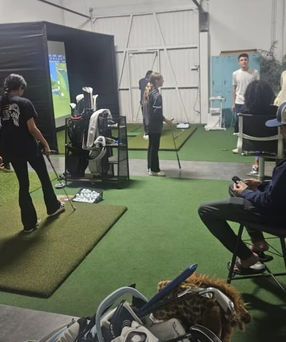 Indoor golf simulator area with several people practicing swings on green turf and large screens.