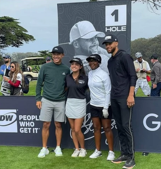 Four people posing on a golf course in front of a large 1 HOLE sign and tournament banner.
