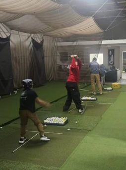People practicing golf swings indoors at a driving range with netted stalls and balls on mats