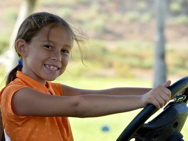 Smiling child in an orange shirt driving a ride-on mower outdoors